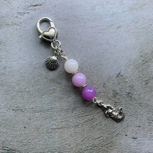 Op de afbeelding: Een zilveren armband met bedels met een hartbedel, een schelpbedel en een zeemeerminbedel. De armband heeft drie kralen in tinten paars en wit.