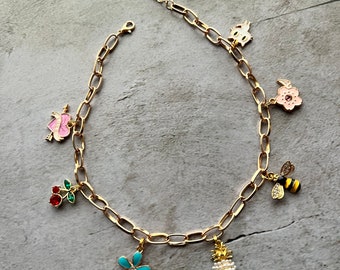 May include: A gold chain necklace with multiple charms, including a pineapple, a flower, a bee, a heart, a cherry, and a flower. The charms are all colourful and detailed.
