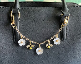 May include: A black handbag with a gold chain and charms. The charms are white flowers and bees.