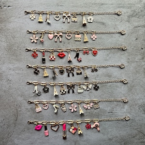 May include: Gold-toned charm bracelets with various miniature charms, including bows, hearts, purses, and Eiffel Towers. The charms are in shades of pink, red, black, and white. The bracelets are displayed on a gray surface.