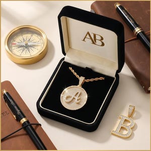 May include: A gold-colored pendant necklace with the letter "A" encrusted in small, sparkling stones, displayed in a black velvet jewelry box. A separate "B" pendant is also visible. A compass and pens are in the background.