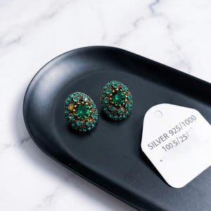 Puede incluir: Un par de pendientes ovalados con una piedra central verde esmeralda rodeada de cristales verdes más pequeños en un marco de metal dorado antiguo. Una etiqueta blanca de joyería tiene el texto SILVER 925/1000 100 5/25 impreso.