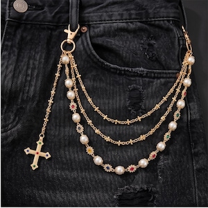 May include: Gold-tone chain accessory with a cross pendant and multiple strands. The chain features faux pearls and colorful gemstone accents. The cross pendant has a colorful gemstone design. The accessory is attached to a pair of black denim jeans.