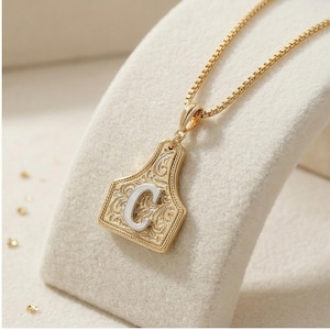 May include: Gold-toned pendant necklace featuring a decorative tag design with the letter "C" in white. The pendant has an ornate, engraved pattern. The necklace chain is a delicate box chain style.