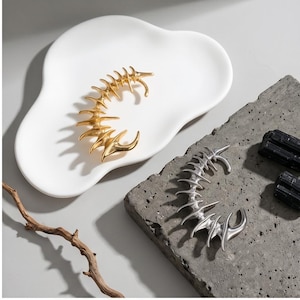 May include: A gold-toned and a silver-toned ear cuff with a unique, spiky design, displayed on a white, organic-shaped dish and a gray textured surface. The jewelry resembles a stylized spine or bone structure. Two black stones are also present.