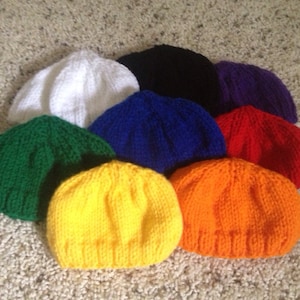 May include: A collection of eight knitted beanies in various colors: white, black, purple, red, blue, green, yellow, and orange. The beanies are arranged in a circular pattern, showcasing their vibrant hues and simple design. The hats are made of a soft, textured yarn.
