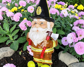 May include: A garden gnome figurine dressed as a firefighter, wearing a black helmet with a yellow Fire Chief 1 emblem, a brown jacket with yellow reflective stripes, and holding a red-handled axe with a silver blade in its hands.