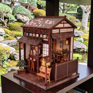 以下が含まれることがあります： 茶色の木材で作られた、瓦屋根と桜の装飾が施されたミニチュアの日本の茶屋モデル。小さなベンチ、盆栽、提灯があり、日本語の「生 の 茶」の文字が見えます。