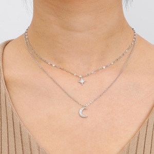 Può includere: Una collana argentata a più strati con un ciondolo a forma di stella e mezzaluna. Lo strato superiore ha una catena delicata, mentre lo strato inferiore mostra un ciondolo a mezzaluna ornato da piccole pietre scintillanti.
