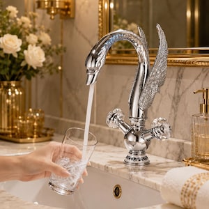 May include: A chrome swan-shaped tap with crystal detailing. Water pours into a clear glass. The tap features a swan head and wing-shaped elements. The bathroom setting includes a white sink and gold-toned accessories.