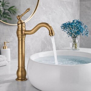May include: A brass-coloured tap with a curved spout, water flowing into a white, round basin. The tap has a vintage design. A round mirror and a vase of blue flowers are in the background. The basin is on a marble countertop.