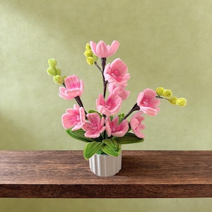 May include: A handcrafted floral arrangement featuring pink blossoms and green foliage in a white, ribbed ceramic pot. The flowers have delicate petals and are arranged on dark stems. The background is a soft green.
