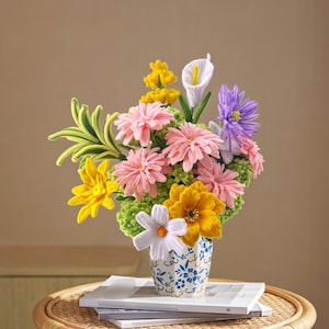 May include: A colorful bouquet of handmade flowers in a small, patterned vase. The arrangement includes pink, yellow, white, and purple flowers, with green foliage. The vase is white with a blue floral design, sitting on a stack of books.