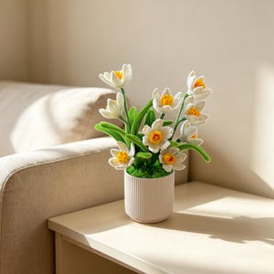 May include: A potted arrangement of white and yellow crocheted daffodil flowers with green stems and leaves. The flowers are planted in a small, cylindrical, off-white ceramic pot featuring a vertical ribbed texture and a green mossy base.
