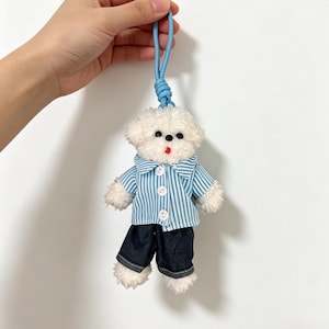 May include: A plush white dog toy with black button eyes, a red mouth, and a blue cord for hanging. The dog is wearing a blue and white striped shirt and dark blue pants.