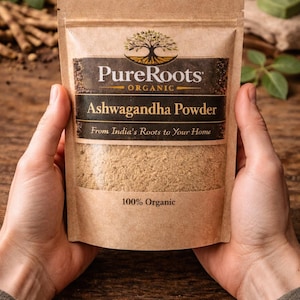 May include: A package of PureRoots Organic Ashwagandha Powder held by hands. The package is a light brown color with a dark brown label. The label includes the text "Ashwagandha Powder" and "From India's Roots to Your Home". The package is labeled "100% Organic".