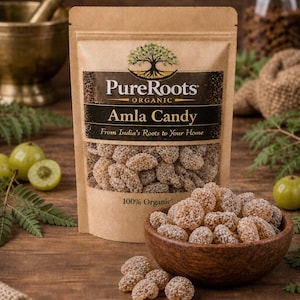 Könnte beinhalten: Eine braune Papiertüte mit "PureRoots Organic Amla Candy" mit einem Baumlogo und eine Holzschale voller Süßigkeiten. Die Süßigkeiten sind hellbraun und körnig. Grüne Früchte und Farnblätter sind um die Schüssel und die Tüte auf einer Holzoberfläche verstreut.