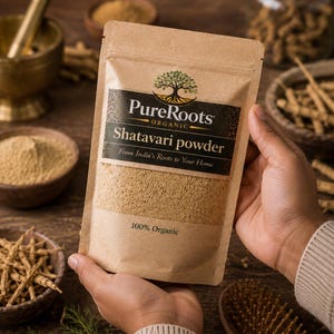 May include: A tan paper bag of Shatavari powder from PureRoots Organic, held in hands. The bag has a black label with the brand name and product description. The words "100% Organic" are printed on the bag. Other bowls and jars of herbs are in the background.