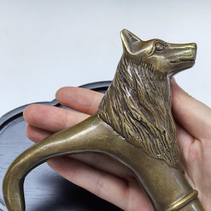 May include: A metallic bronze-toned walking stick handle featuring a sculpted wolf head with detailed fur texture and a curved grip. The handle includes a cylindrical base attachment point, showcasing a polished finish with dark patina accents in the crevices.