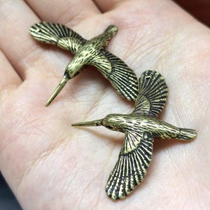 May include: Two small, metallic, gold-toned bird charms with detailed engraved wing feathers and long, pointed beaks. The birds are depicted in a flying position with wings spread wide, showcasing a textured, antique-style finish on the metal surface.