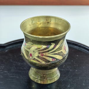 May include: A small brass cup with a flared rim and a pedestal base. The exterior features a dark, tarnished finish with etched floral patterns highlighted in gold and small red accents. The interior of the cup shows a polished brass surface.