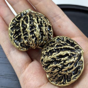 May include: Two metallic objects shaped like walnut shells with a textured, uneven surface. The items feature a dark, oxidized finish within the deep crevices and a polished, bright gold-toned finish on the raised ridges of the shell exterior.