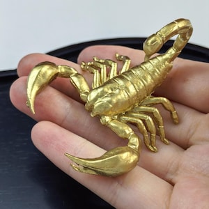 Op de afbeelding: Een kleine, metallic goudkleurige schorpioenfiguur met een gesegmenteerd lichaam, acht gelede poten, twee grote voorste scharen en een gebogen staart met een angel. Het oppervlak heeft een gestructureerde, matte gouden afwerking met details.