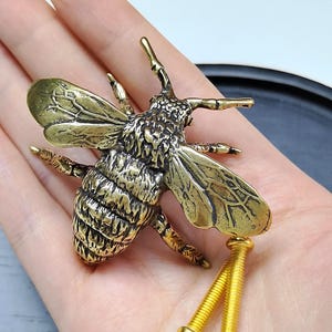 May include: A brass bee-shaped cabinet knob with detailed wings and a textured body. The bee features two long, gold-toned metal screws extending from the underside for mounting purposes, showcasing a metallic finish and intricate insect anatomy.