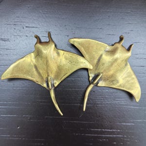 Op de afbeelding: Twee messingkleurige metalen ornamenten in de vorm van mantaroggen. Elk stuk heeft een gladde, metalen afwerking met een puntige staart, brede borstvinnen en twee kleine koplobben bij de kop. De figuren zijn in een platte houding.