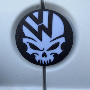 May include: A circular emblem featuring a black textured border and a white skull graphic integrated with a stylised letter W. The design combines a skull face with sharp, angular features and a prominent W shape at the top, creating a high-contrast look.