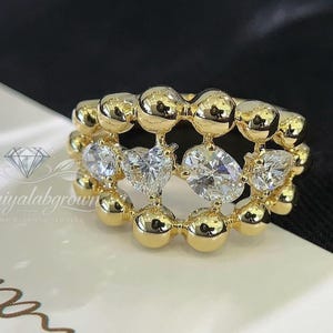May include: A gold ring with a distinctive design. The ring incorporates small, round gold beads and several clear, sparkling gemstones, including heart-shaped and oval-cut stones. The ring is set against a white and black background.