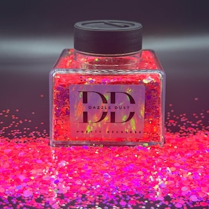 May include: A clear, square jar filled with vibrant pink glitter, topped with a black lid. The label reads "DD DAZZLE DUST PRETTY RECKLESS". Glitter is scattered around the jar, creating a sparkling effect. The background is a dark gradient.