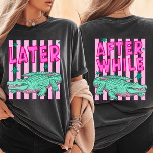 May include: Dark gray t-shirt with a front and back design. The front says "LATER" in pink letters with a green alligator graphic. The back says "AFTER WHILE" in pink letters with a green alligator graphic.