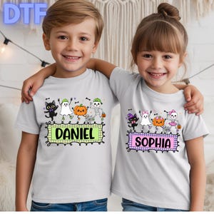 May include: Two light gray short-sleeve t-shirts featuring Halloween-themed graphics of a black cat, a ghost, a pumpkin, and a skeleton. The left shirt displays the name DANIEL in a green box, while the right shirt displays the name SOPHIA in a pink box.