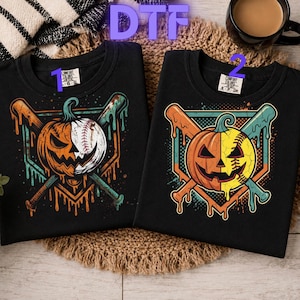 May include: Two black t-shirts featuring a graphic of a pumpkin split with a baseball or softball, crossed bats, and a home plate. The design includes dripping paint effects in orange and teal. The shirts have a Comfort Colors label at the neck.