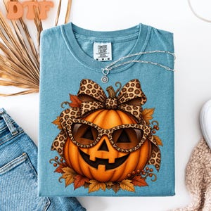 May include: A blue t-shirt featuring a graphic of an orange jack-o'-lantern wearing leopard print sunglasses and a matching leopard print bow. The pumpkin is surrounded by autumn leaves and berries. A silver chain necklace rests on the shirt.