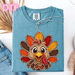 May include: A blue short-sleeve t-shirt featuring a graphic of a turkey with frayed-edge feathers in shades of orange, red, yellow, and brown. The turkey has large eyes and a yellow beak. The shirt has a Comfort Colors label at the neck.