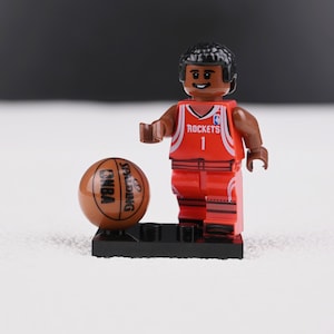 May include: A red and white Lego basketball player figurine with the number 1 on the jersey, and a brown basketball with "NBA" and "Spalding" printed on it. The figurine is standing on a black base.