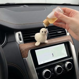 May include: A small, cream-colored needle-felted seal figurine attached to a car air vent. A hand holds a small, clear plastic bottle with a wooden cap, dripping a single drop of light-colored liquid onto the top of the seal's head.