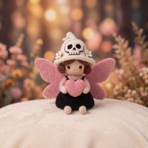 May include: A needle-felted doll featuring a cream-colored hat with a black skull motif and small white flowers. The doll has brown hair, pink wings, and a black dress, while holding a pink heart in its hands. The figure has a soft texture.