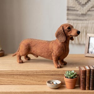 Op de afbeelding: Een naaldgevilte bruine teckel-figuur die in profiel staat. De hond heeft een textuurrijke, pluizige vacht, lange hangende oren en een zwarte neus. Het is gemaakt met realistische verhoudingen, waarbij het langgerekte lichaam is vastgelegd.