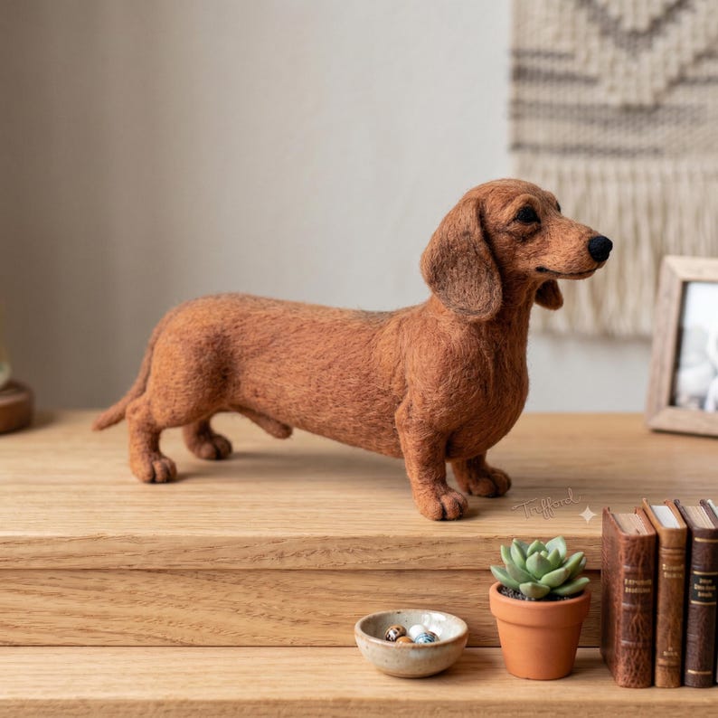 Op de afbeelding: Een naaldgevilte bruine teckel-figuur die in profiel staat. De hond heeft een textuurrijke, pluizige vacht, lange hangende oren en een zwarte neus. Het is gemaakt met realistische verhoudingen, waarbij het langgerekte lichaam is vastgelegd.