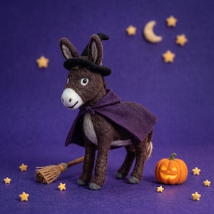 May include: A needle-felted brown donkey figurine wearing a black witch hat and a purple cape. The donkey stands next to a small wooden broomstick and a miniature orange jack-o'-lantern pumpkin, surrounded by small yellow felt stars.