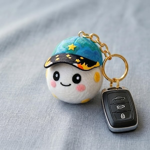 Puede incluir: Un llavero caprichoso con un personaje blanco y redondo con una cara sonriente, que lleva una gorra azul adornada con estrellas amarillas. El llavero tiene una cadena y un anillo dorados, unidos a un mando de coche negro.