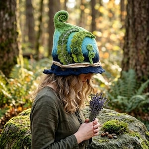 May include: A felted hat featuring a gradient of blue and green colors, a curled top, and decorative fern-like leaf appliqués. The hat includes a braided cord trim around the base and a wide, wavy brim, showcasing a textured, handcrafted appearance.