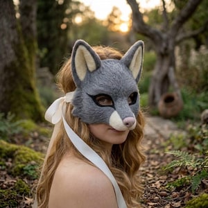 May include: A gray felted animal mask featuring pointed ears with cream-colored inner details, black eye markings, and a light-colored snout with a pink nose. The mask is secured with a long, smooth white ribbon tied at the side of the head.