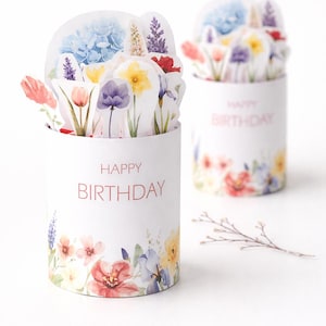 Peut inclure: Deux contenants cylindriques blancs avec des motifs floraux et l'inscription "HAPPY BIRTHDAY" en rouge. Les contenants sont remplis de découpes de papier en forme de fleurs. Les découpes représentent diverses fleurs comme des tulipes, des iris et des hortensias.