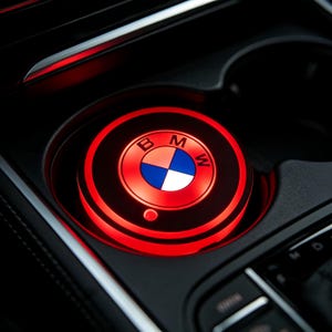 Puede incluir: Un posavasos con el logotipo de BMW iluminado en rojo en un portavasos de coche. El posavasos tiene un diseño circular con el emblema de BMW en el centro, con segmentos azules, blancos y naranjas. El interior del coche es negro.