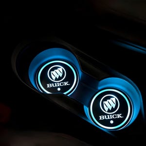 May include: Two illuminated Buick car cup holder inserts. Each insert features the Buick logo in white against a black background, surrounded by a bright blue light. The inserts are set in a black car console.