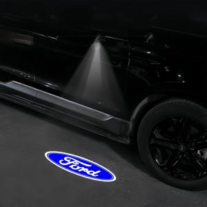 May include: A black car with a blue and white Ford logo projected onto the ground. The logo is an oval shape with the word "Ford" in a stylized font. The car's door is open, and a light shines down.